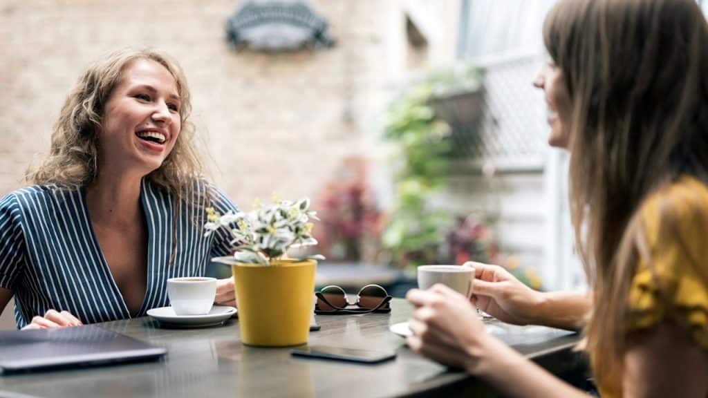 Two women are sitting at an outdoor table with coffee, and one woman is laughing while looking at the other.