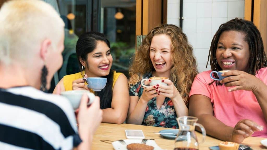 A group of women laugh together at a cafe while holding coffee cups.