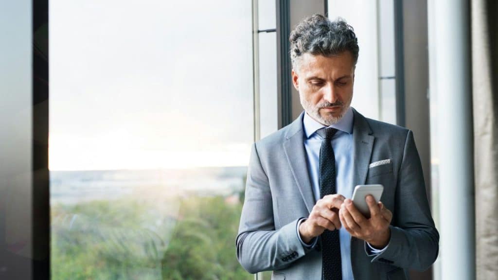 A serious, well-dressed man stands by a window and looks at his smartphone.