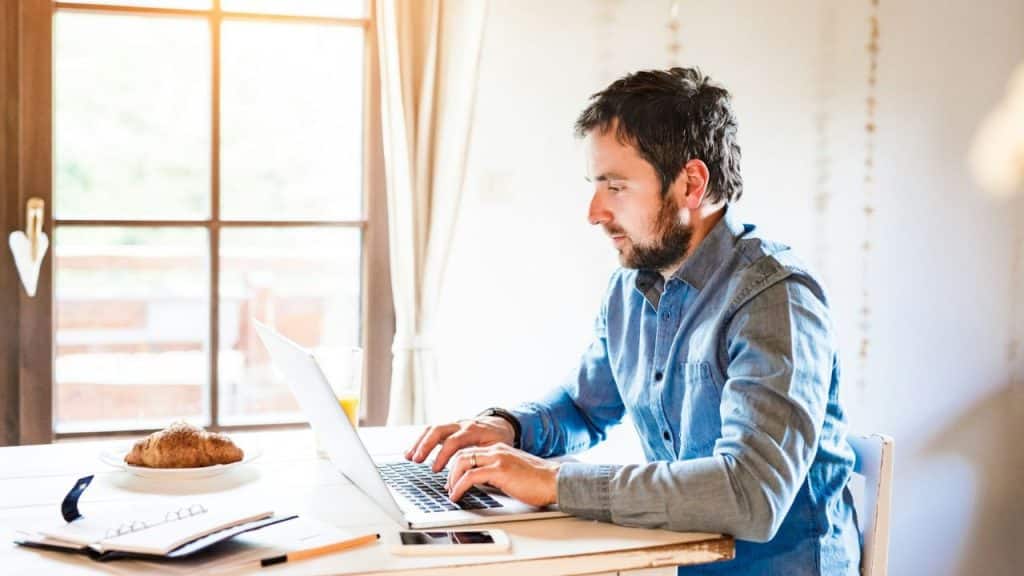 A bearded man is intently working on a laptop at a table in a brightly lit room.