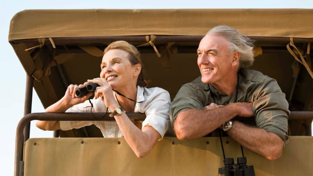 A smiling older couple is on a safari vehicle, with the woman holding binoculars.
