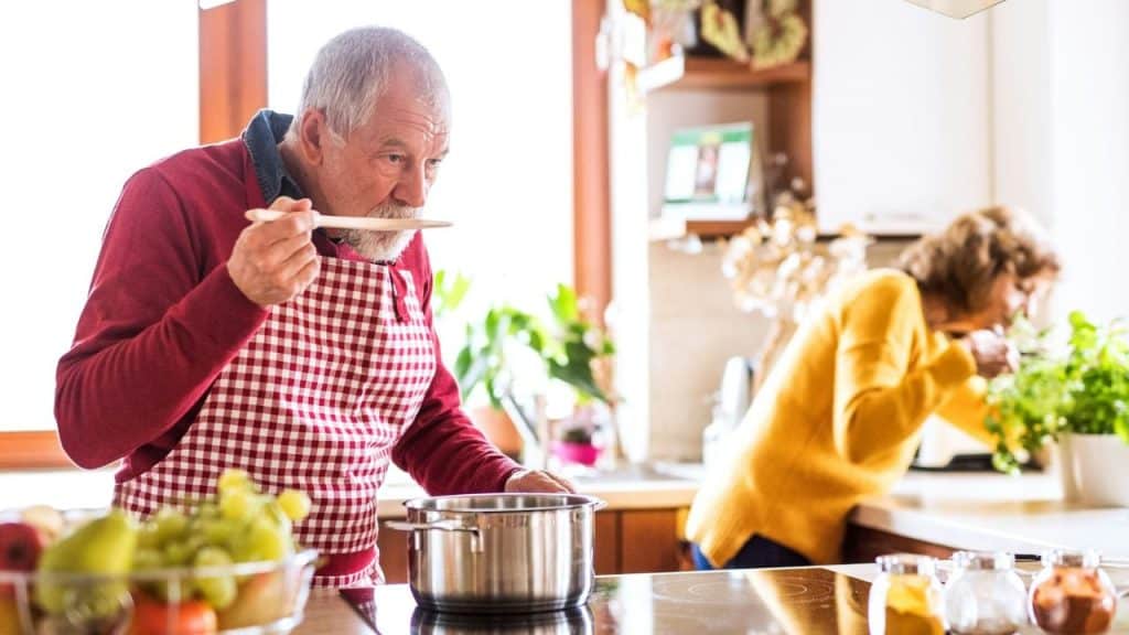 An elderly man tastes food from a pot while a woman sniffs a plant.