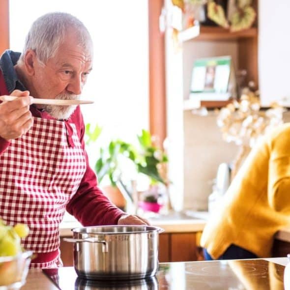 An elderly man tastes food from a pot while a woman sniffs a plant.