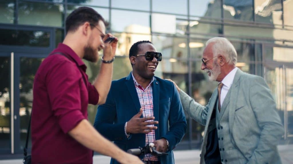 Three men of different ages and ethnicities are laughing and talking outdoors in front of a glass building.