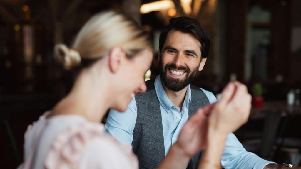 A bearded man smiling while talking with a woman at a restaurant table.