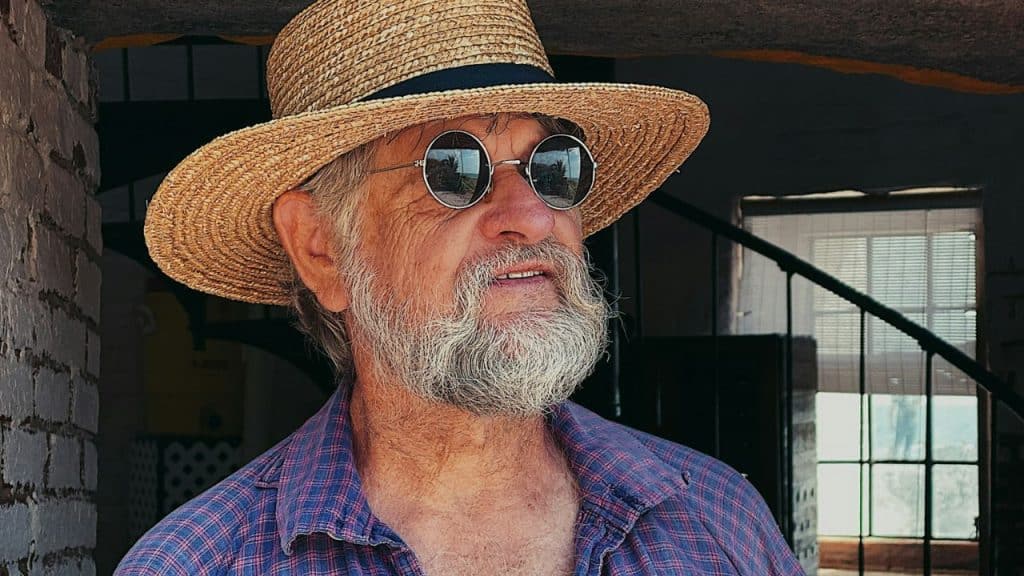 A man with a white beard, a straw hat, and round sunglasses looks to the side.
