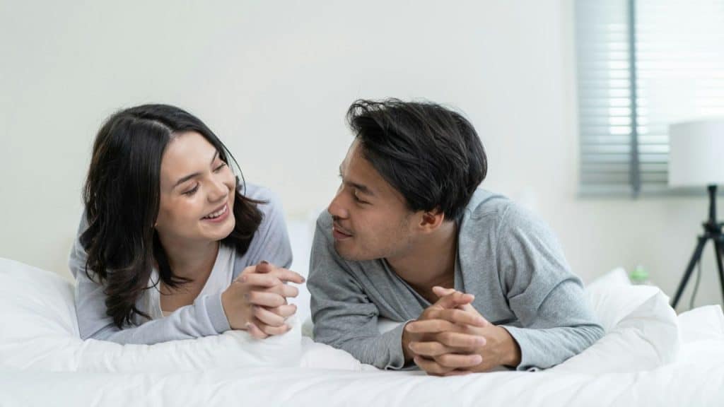 A happy man and woman are lying on a bed, looking at each other and smiling.