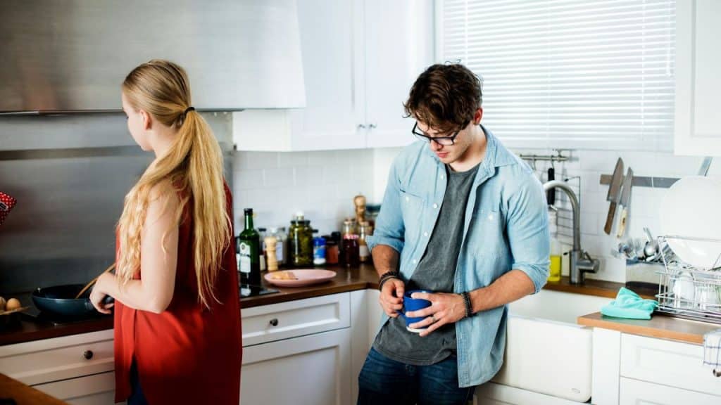 A couple in a kitchen. The woman is cooking while the man drinks coffee.