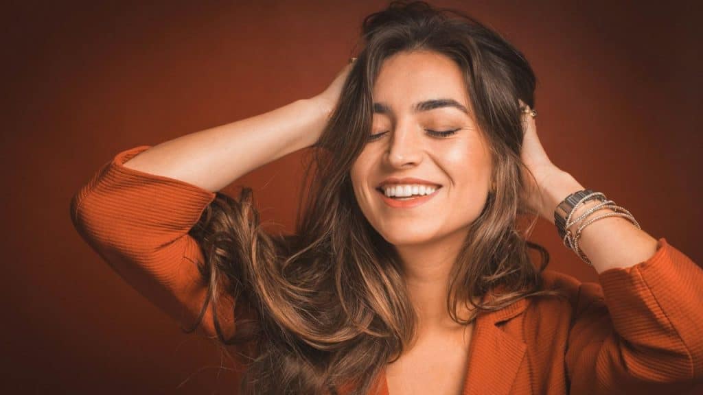 A happy woman in a rust-colored jacket closes her eyes and smiles while holding her hands in her hair.
