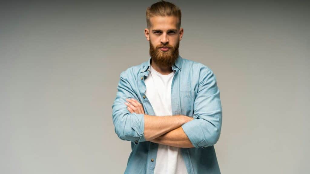 A man with a full beard and a light-colored shirt stands with his arms crossed