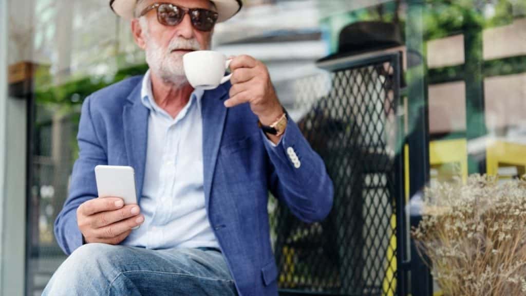 A stylish older man sips coffee while using his phone outdoors.