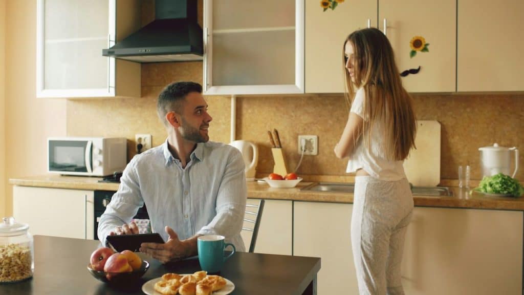 A man is sitting at a kitchen table with a tablet, looking at a woman who is standing and looking at him.