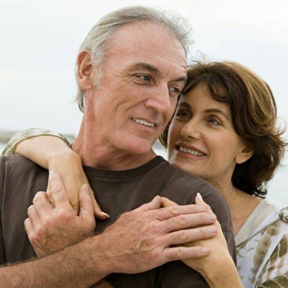 A happy older couple embraces on a beach, looking at each other with affection.