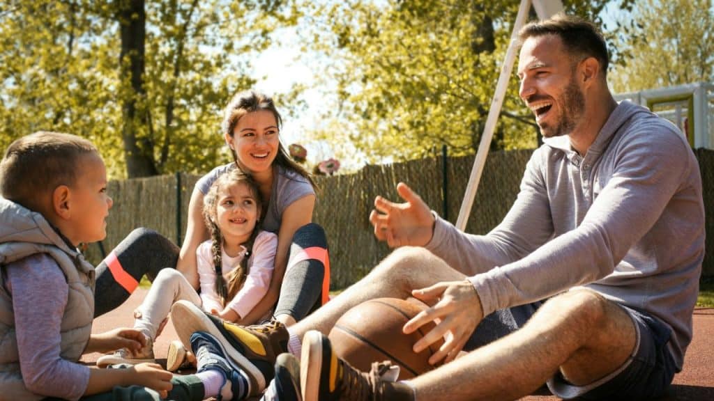 A happy family with two young children sits outside with a basketball on a sunny day.