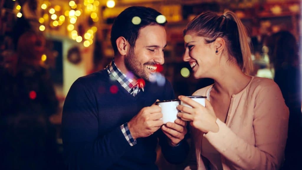 A man and woman are smiling and clinking white mugs in a warmly lit, festive setting.