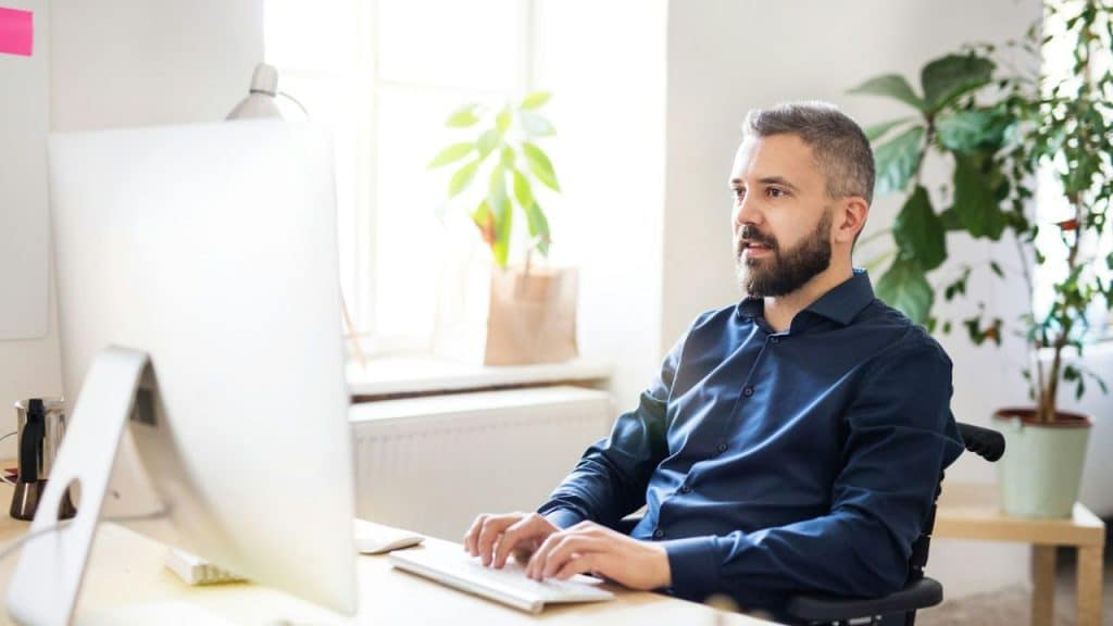 A focused man with a beard sits at a desk working on a computer in a bright office.