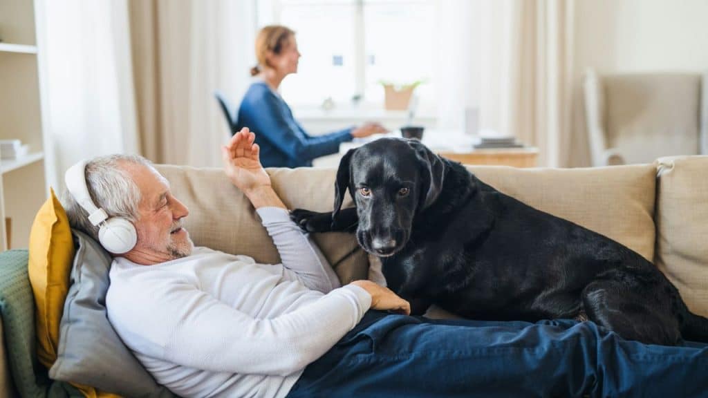 An elderly man with headphones relaxes on a couch with a black dog.