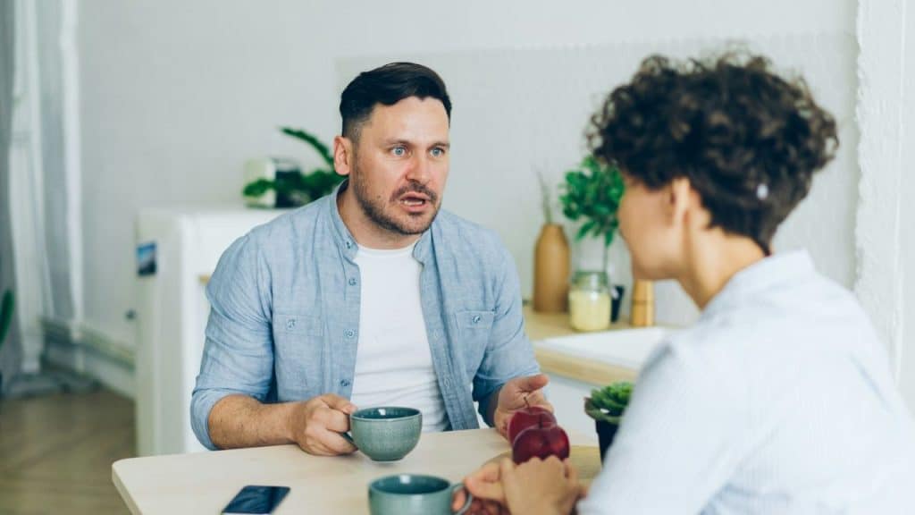 A man and woman sit at a table; the man is speaking with an animated, open-mouthed expression.