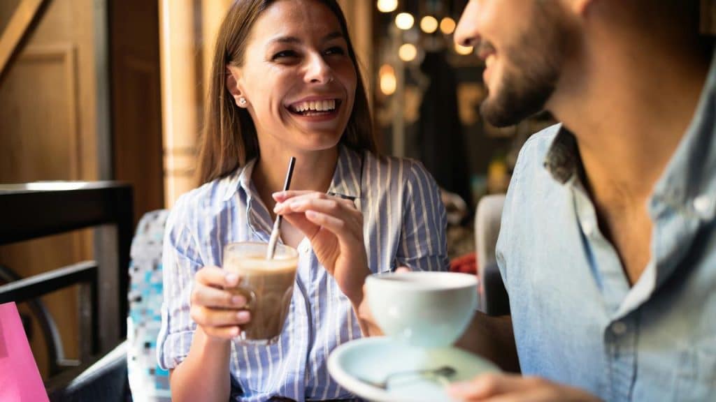 A smiling woman holding an iced coffee while talking with a man at a café.