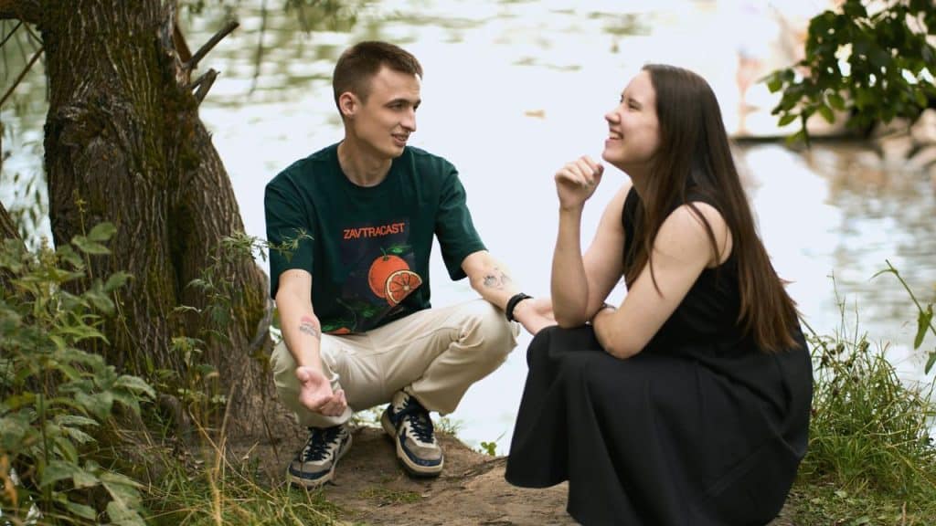 A smiling man in a green shirt crouches and talks to a woman by a river.