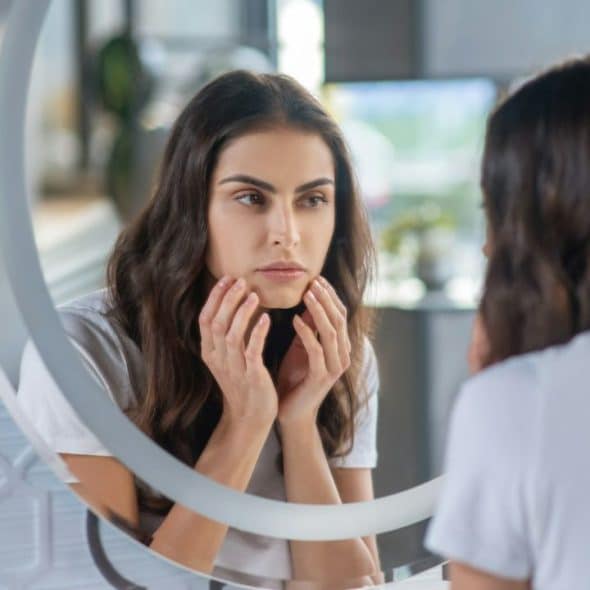 A woman with her hands on her face looks at her reflection in a round mirror.