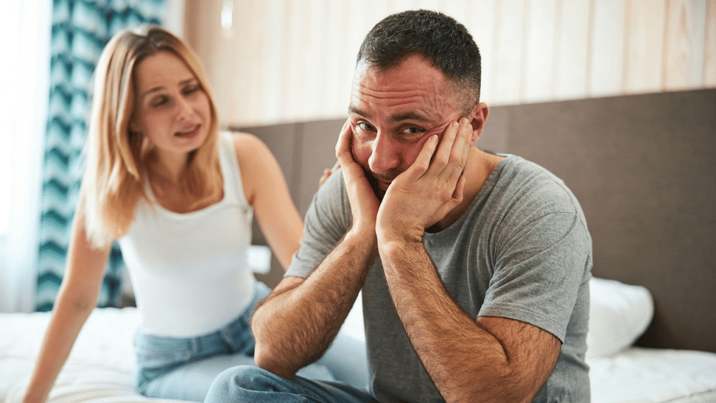 A distraught man with his face in his hands looks at the camera while a woman sits behind him.