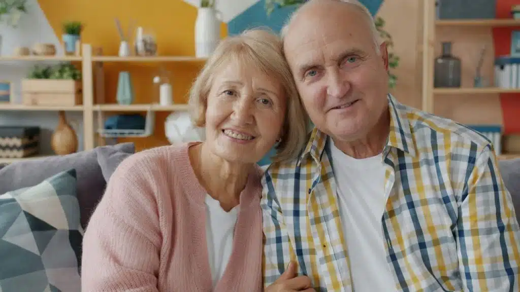 Smiling older couple, woman in pink cardigan and man in plaid shirt, seated indoors.