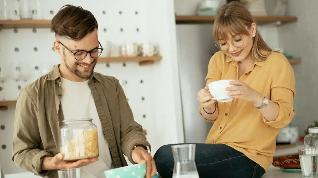 Couple smiling in a bright kitchen, man holding a jar, woman with a white mug.