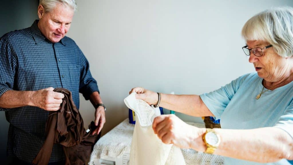 An older man and woman are indoors, folding clothes together on a table.