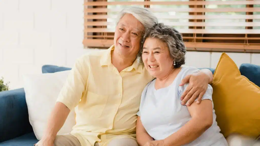 Smiling elderly Asian couple seated close together on a blue couch, embracing indoors.