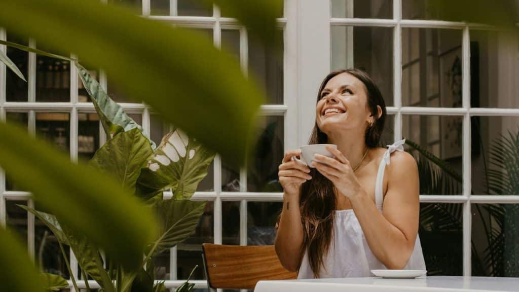 Smiling woman in a white top holding a teacup, looking up near a window.