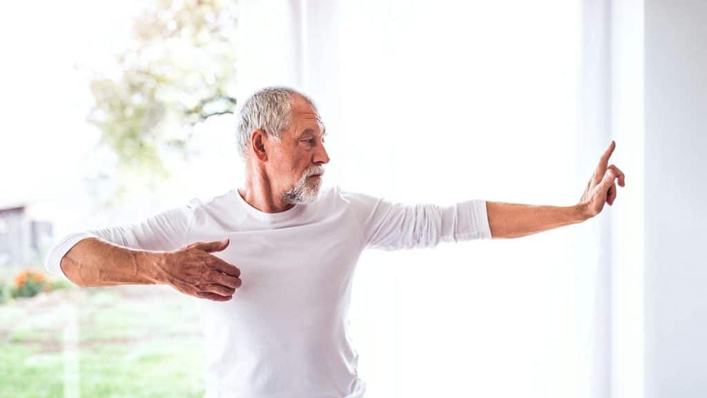 Older man in a white long-sleeved shirt doing a gentle exercise indoors near a window.