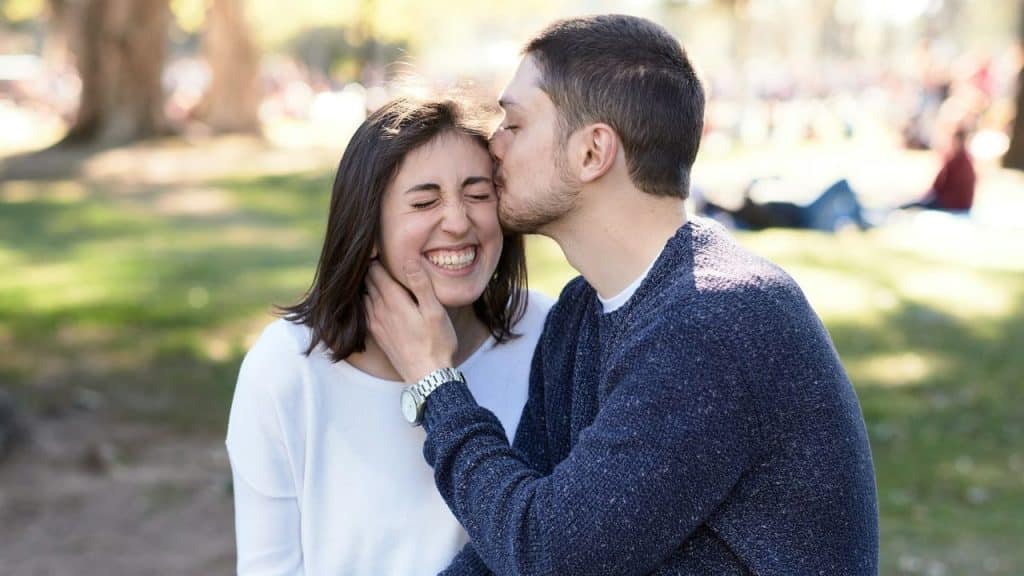 Young man in a navy sweater kissing a smiling woman on the forehead outdoors.