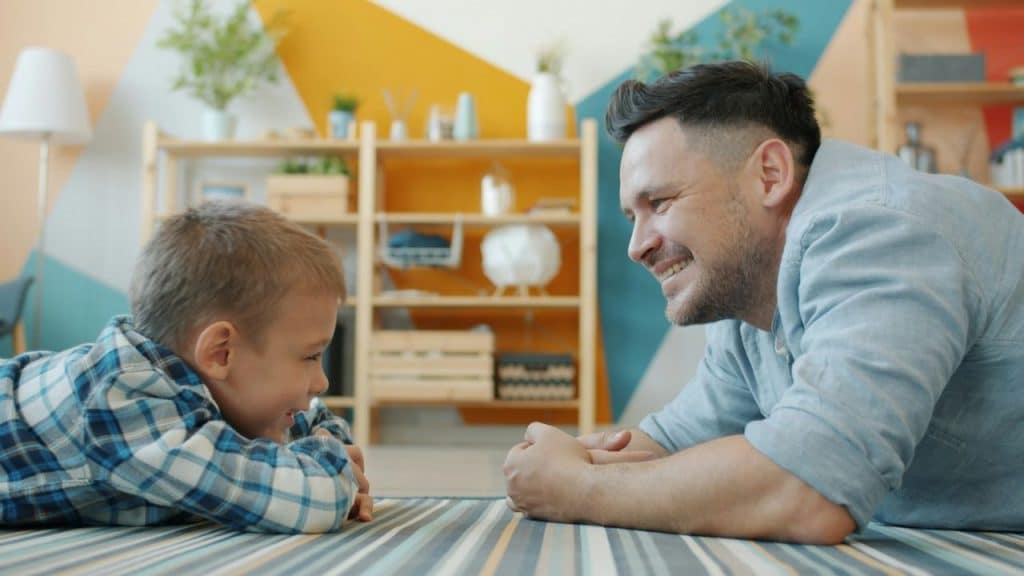 Father and young son lying on the floor, smiling at each other indoors.