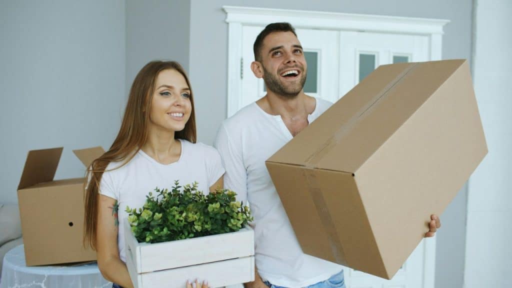 Smiling couple carrying moving items, the man with a box, the woman with a plant box.