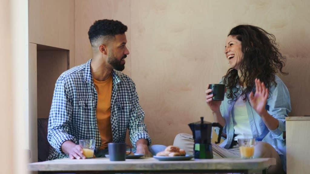 Smiling couple sitting at a wooden table, drinking coffee and laughing indoors.