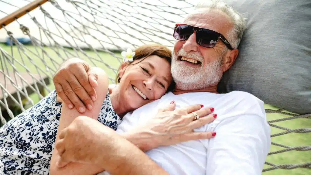 Smiling elderly couple hugging in a hammock, the man wearing sunglasses and a beard.