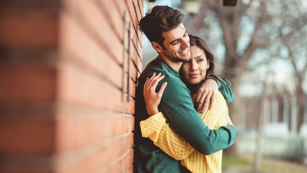 Smiling man and woman embracing each other outdoors beside a brick building.