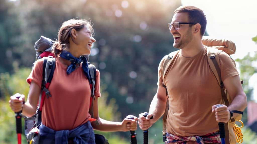 Smiling couple wearing backpacks and holding trekking poles, looking at each other outdoors.