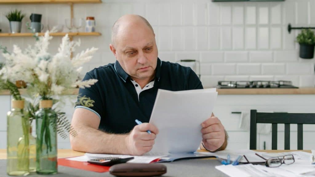 A concerned, middle-aged man reads a document intently at a kitchen table.