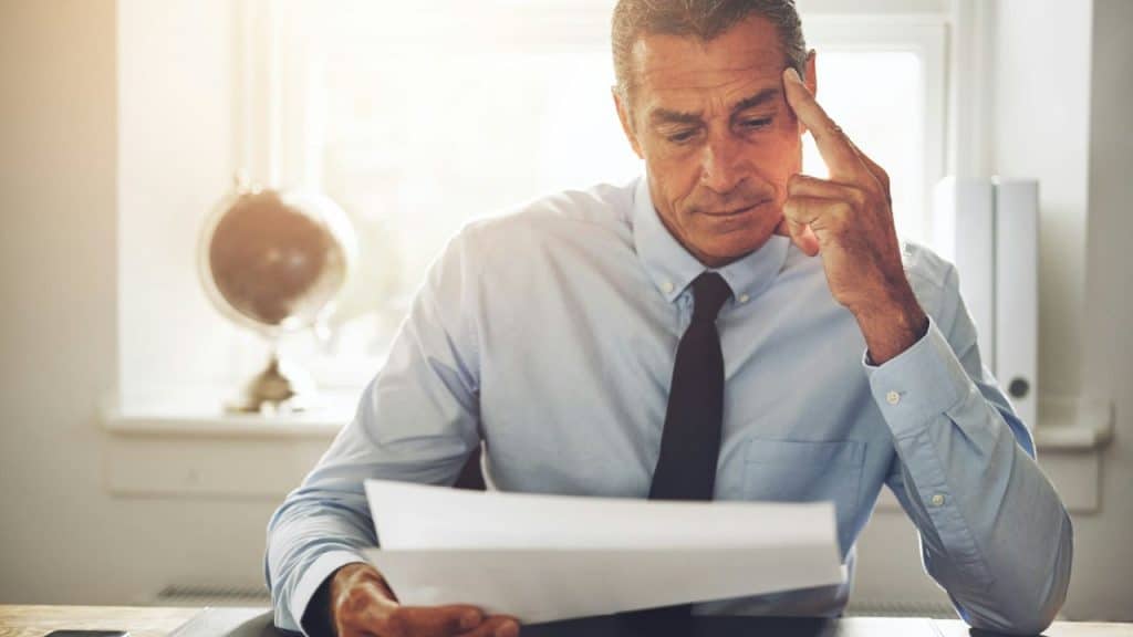 A focused man in a dress shirt reads a paper at his desk, deep in thought.