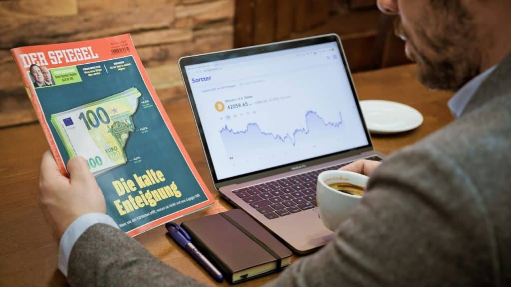 A man at a table reviews a financial chart on a laptop, holding a magazine and coffee.