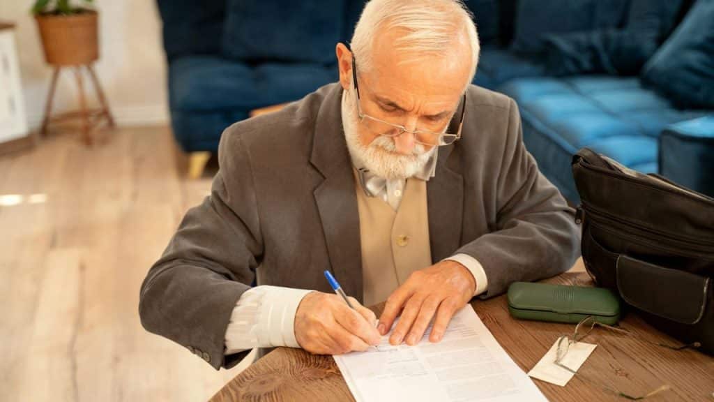A distinguished older man with a white beard signs a legal document at a table.