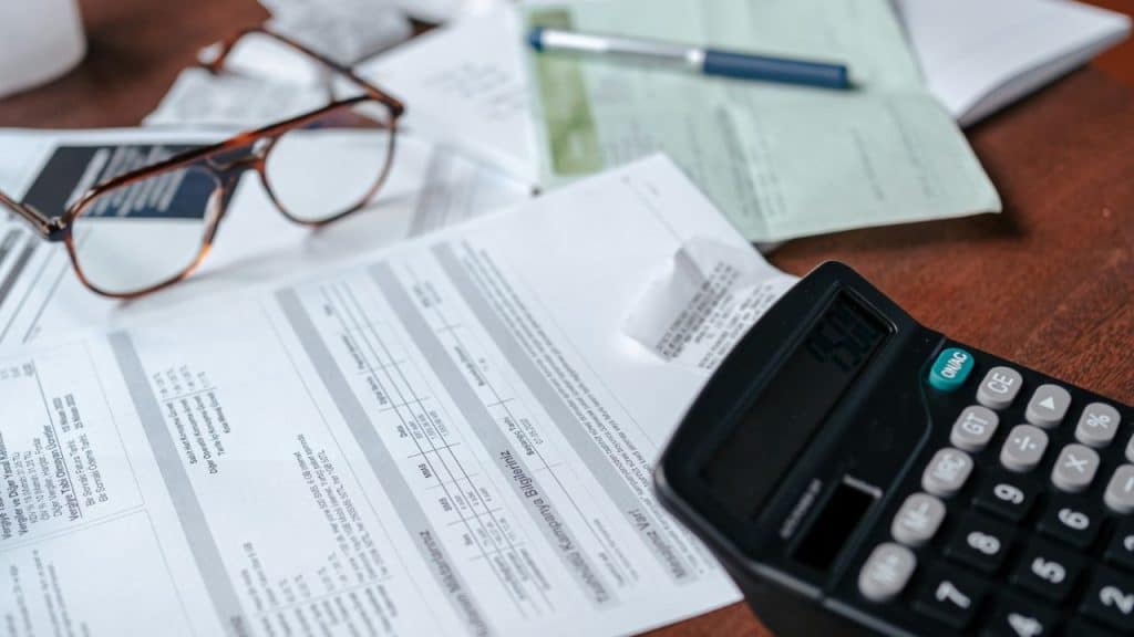 Eyeglasses, a calculator, bills, and papers are scattered on a wooden table.