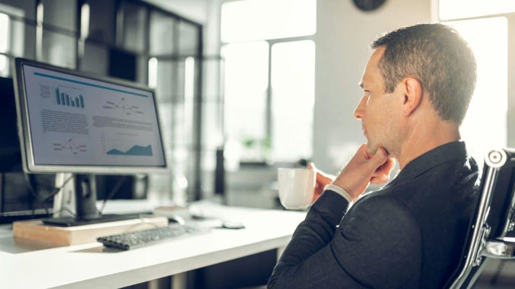 A man in a suit sips coffee while studying financial graphs on a bright computer monitor.