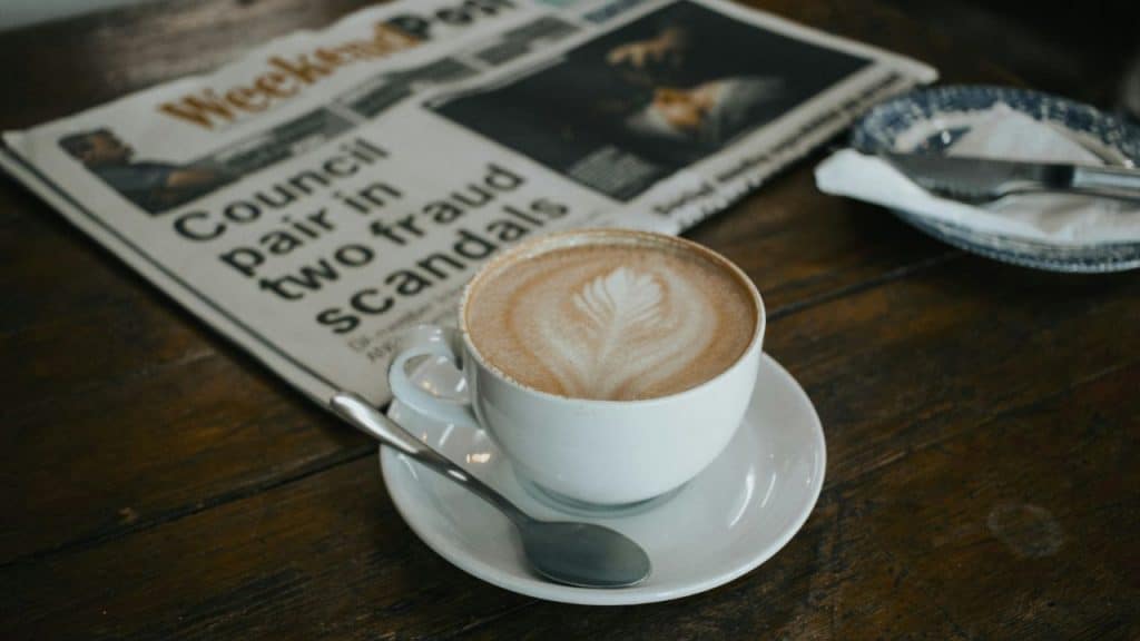 A cappuccino with foam art sits on a saucer next to a folded newspaper.