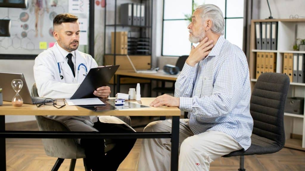 A male doctor reviews a clipboard while a senior male patient touches his throat.