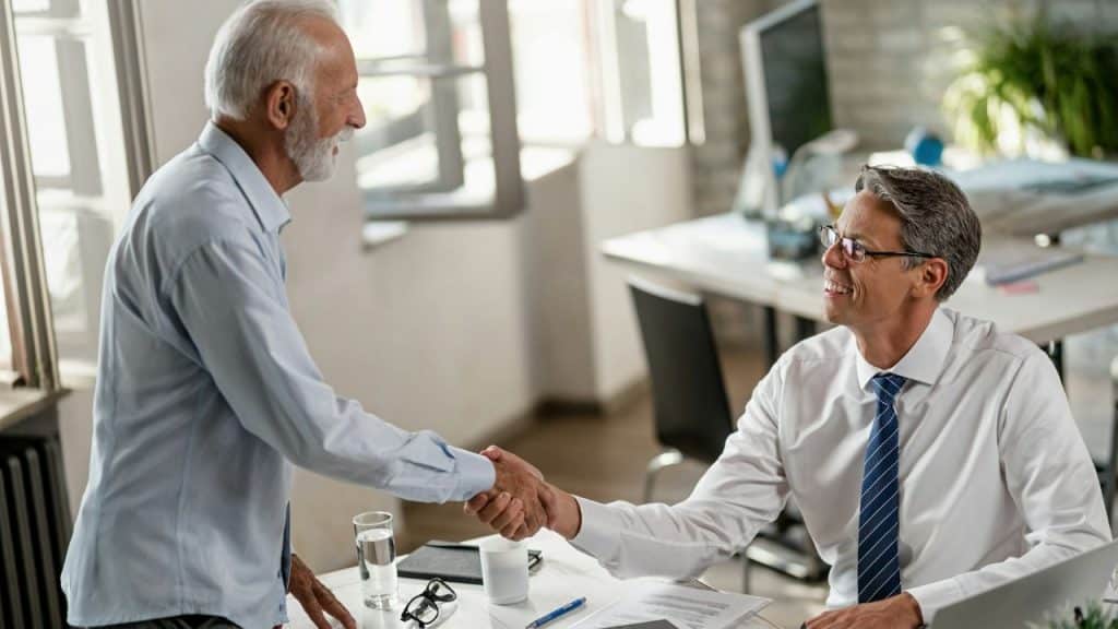 Two professional men, one older and one middle-aged, happily shake hands across a desk.