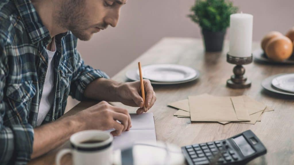 A focused man in a plaid shirt writes with a pencil at a wooden dining table.