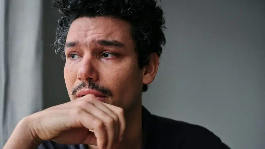 A concerned man with curly hair and a mustache looks away, resting his chin on his hand.